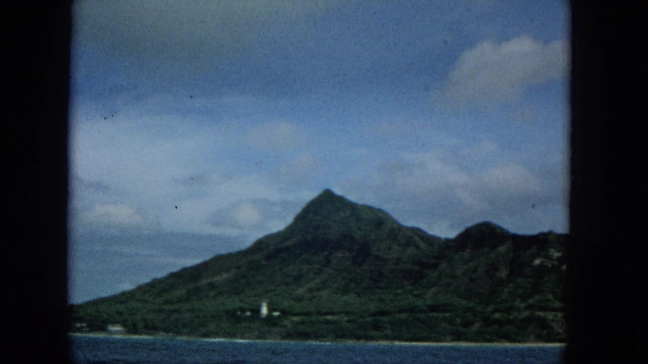 1965 8mm boat-view frame of Molokai with a steep green mountain, blue water, partly cloudy sky, and a small white tower-like coastal structure.