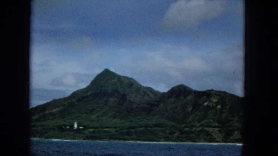 1965 8mm frame of Molokai coastline showing green ridges, a narrow beach or shoreline, blue water, clouds, and a small white tower-like structure near the coast.