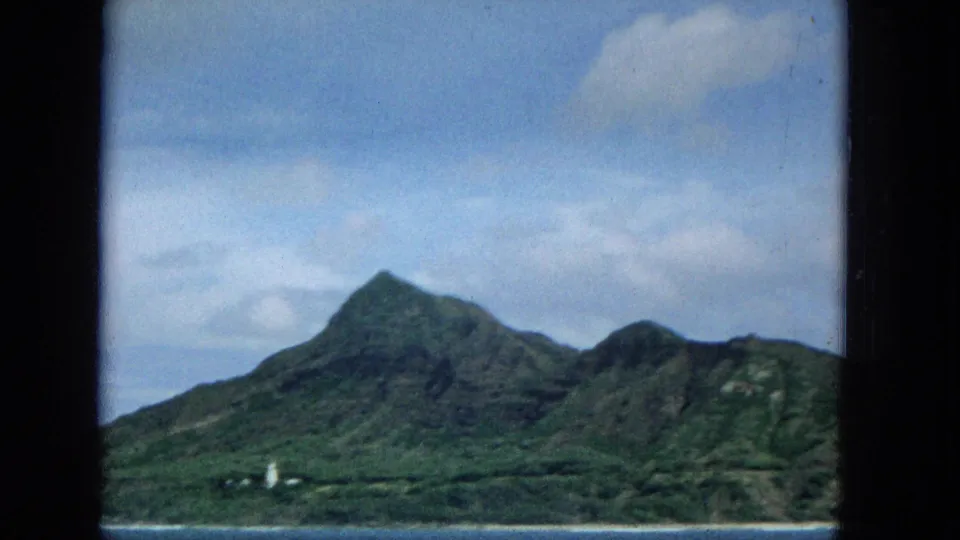 1965 8mm view of Molokai from offshore, with green mountain slopes, blue ocean foreground, light shoreline, clouds, and dark film or window borders.