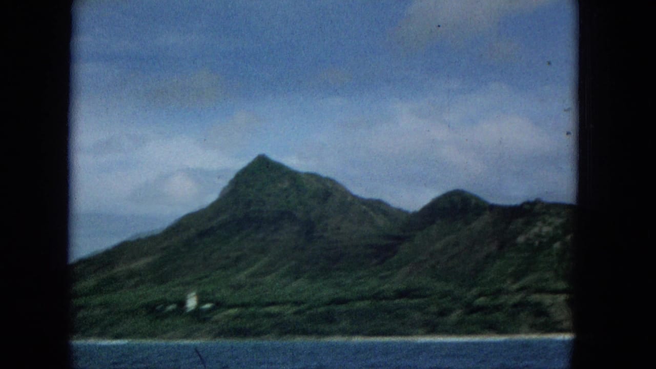 1965 Super 8 frame of Molokai coastline from the water, showing a green mountain, cloudy sky, and small white lighthouse-like structure near shore.