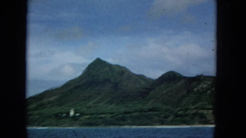 1965 Super 8 frame of Molokai coastline from the water, showing a green mountain, cloudy sky, and small white lighthouse-like structure near shore.