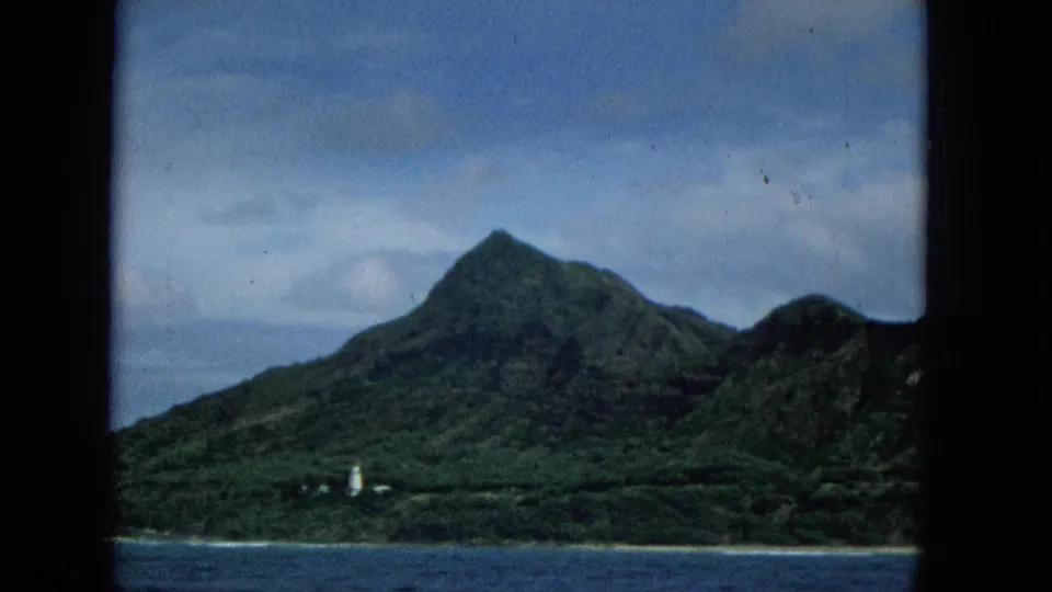 Boat-shot 1965 Super 8 view of the Molokai shore, with a steep green mountain, low coastline, blue water, and a small white lighthouse-like feature.