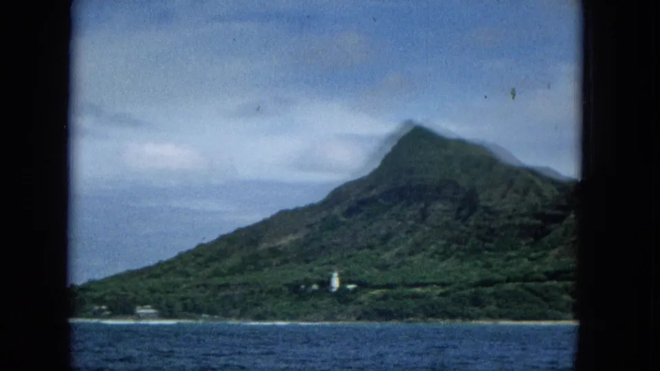 1965 Molokai home-movie frame showing the island from a moving boat, with textured blue water, green slopes, and a white coastal lighthouse-like structure.