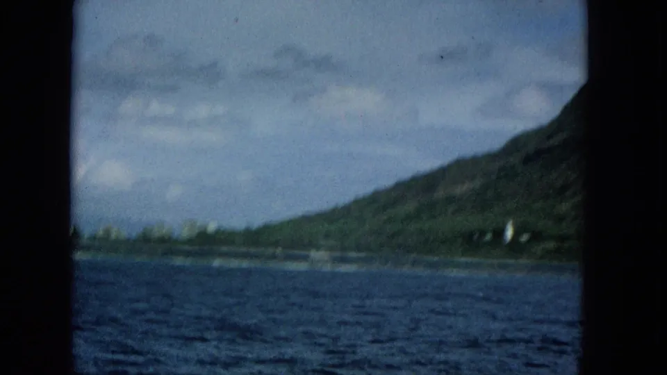 Final 1965 Super 8 frame from the Molokai approach, with dark blue ocean in front, a green shoreline at right, cloudy sky, and small pale coastal structures.