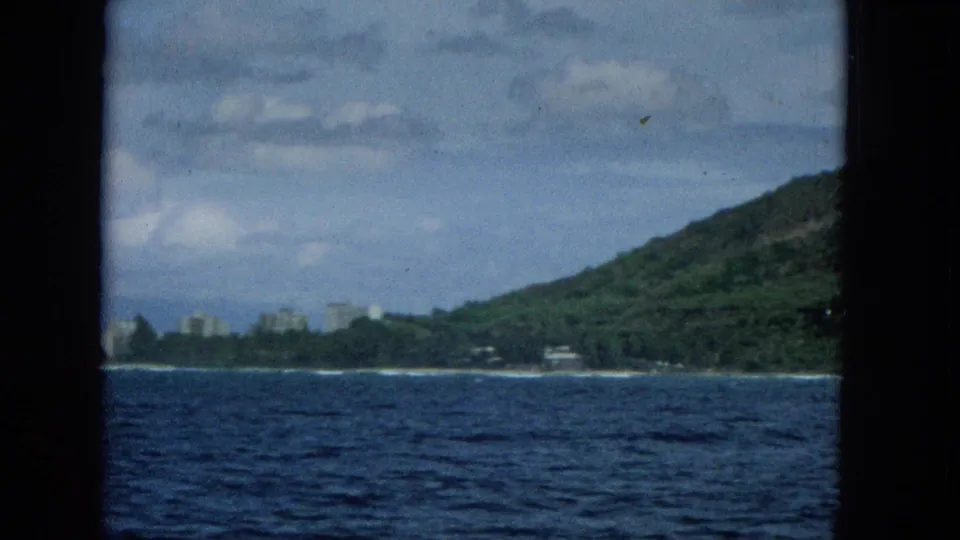 1965 home-movie frame looking from a boat toward a Hawaiian island coastline, with blue water, a green mountain slope, and small distant buildings along the shore.