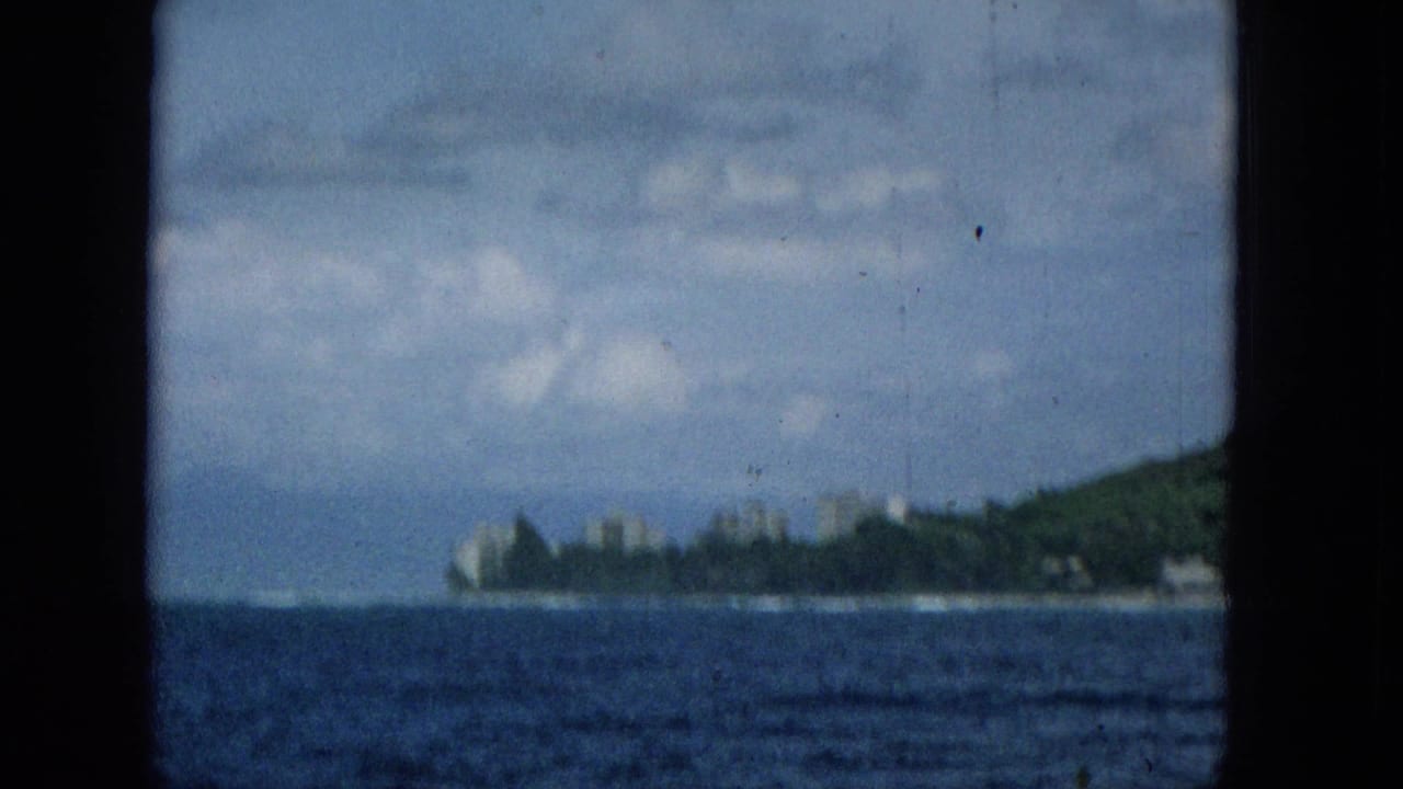 Soft 1965 travel footage frame showing a distant Hawaiian coastline across open blue water, with pale buildings near the shore and dark borders suggesting a boat window or film gate.