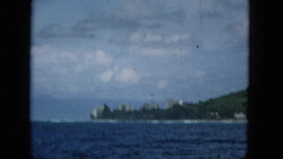 Soft 1965 travel footage frame showing a distant Hawaiian coastline across open blue water, with pale buildings near the shore and dark borders suggesting a boat window or film gate.