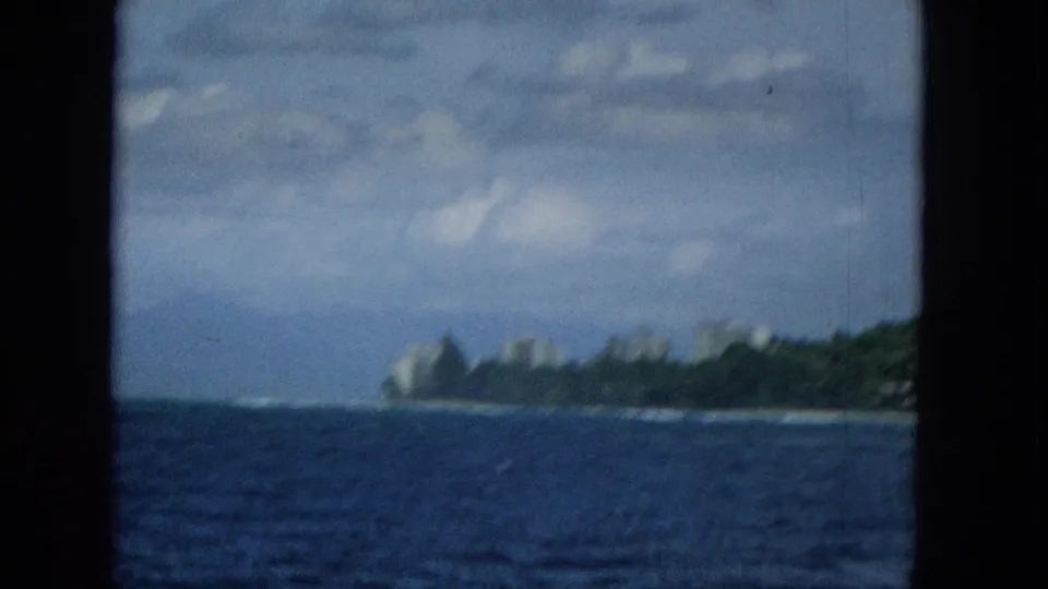 A soft, grainy 1965 home-movie view of a Hawaiian shoreline from the water, with low surf, distant coastal structures, and a cloudy sky above the island edge.