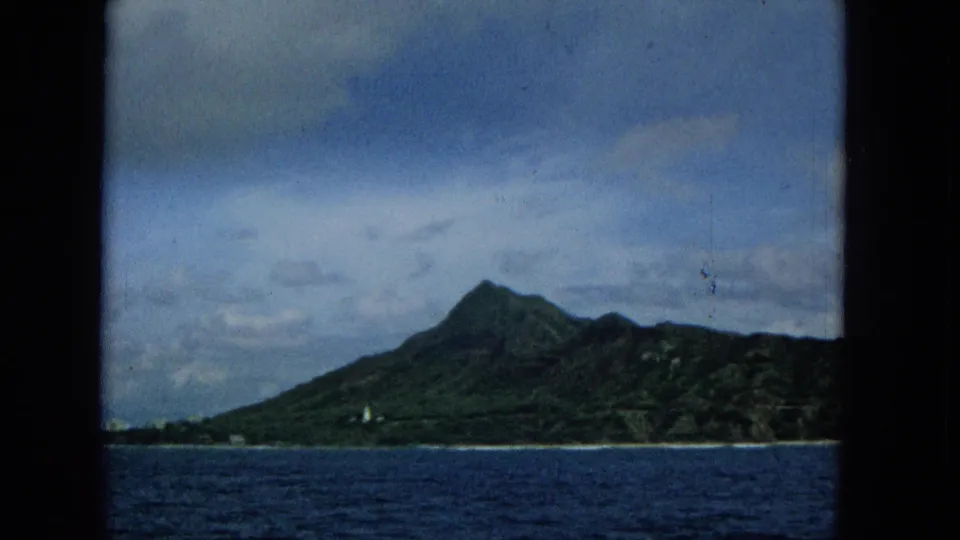 1965 home-movie frame from a boat facing a Hawaiian island mountain ridge, with dark blue water in the foreground and a narrow developed shoreline beneath cloudy skies.