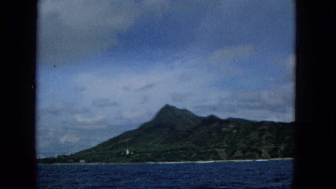 1965 8mm home-movie frame of a Hawaiian island mountain seen from offshore, with ocean foreground, cloudy sky, and dark border at the left edge.