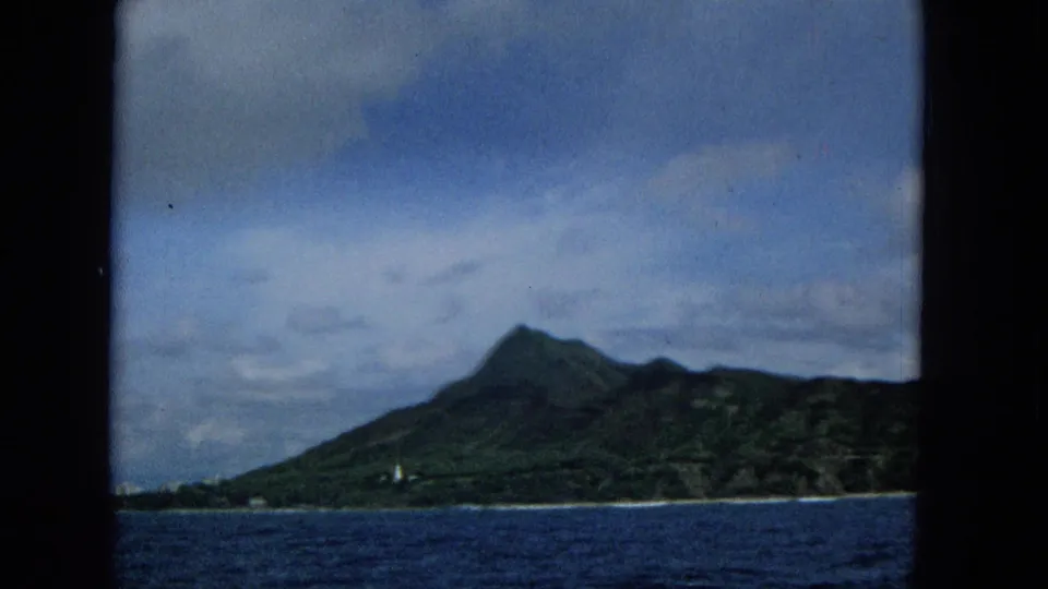 1965 8mm home-movie frame of a Hawaiian island mountain seen from offshore, with ocean foreground, cloudy sky, and dark border at the left edge.