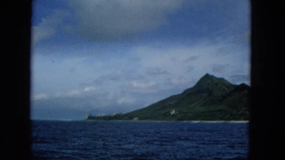 1965 8mm home-movie view of a Hawaiian island mountain and low coastline beyond choppy blue water, framed by dark film or window edges.
