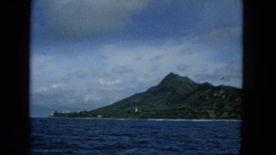 1965 8mm frame of a distant Hawaiian island mountain seen over dark ocean water, with a pale shoreline structure and cloudy sky visible.