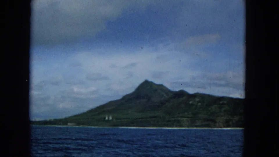 1965 home-movie view of a green Molokai shoreline and pointed mountain seen from offshore across blue water.