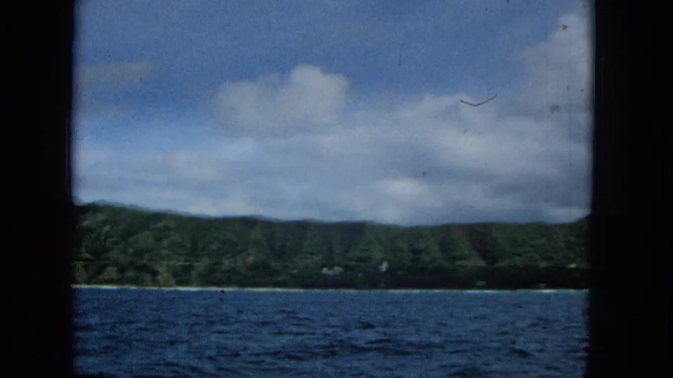 A softer 1965 8mm frame shows the Molokai shoreline with low green hills, blue water, and broad clouds from an offshore viewpoint.
