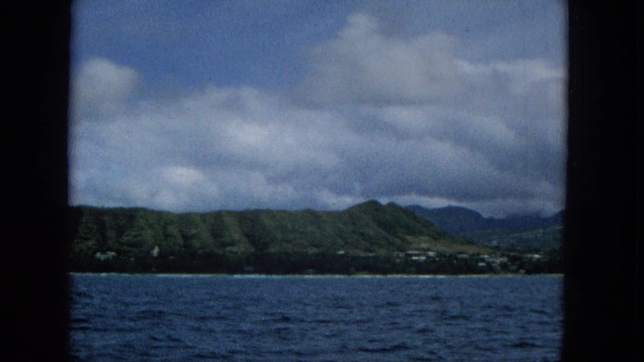 1965 8mm footage shows blue ocean in the foreground with a distant Hawaiian shoreline, green hills, scattered small buildings, and heavy clouds framed by dark boat edges.