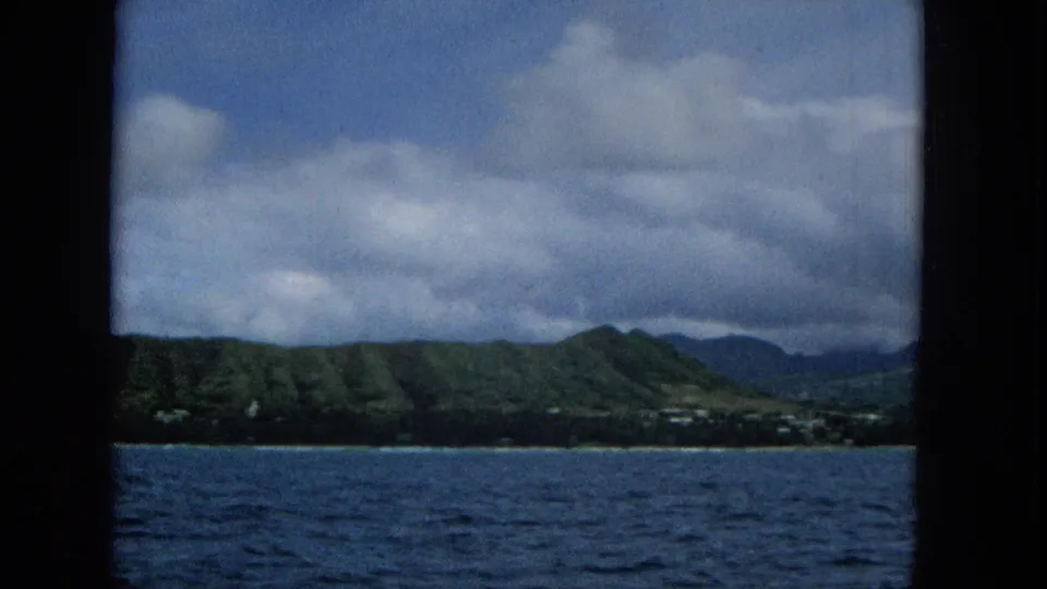 1965 8mm footage shows blue ocean in the foreground with a distant Hawaiian shoreline, green hills, scattered small buildings, and heavy clouds framed by dark boat edges.