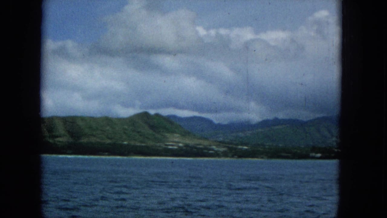 A boat passenger's view looks across the ocean toward green Hawaiian island hills and a low shoreline beneath a cloudy blue-gray sky.