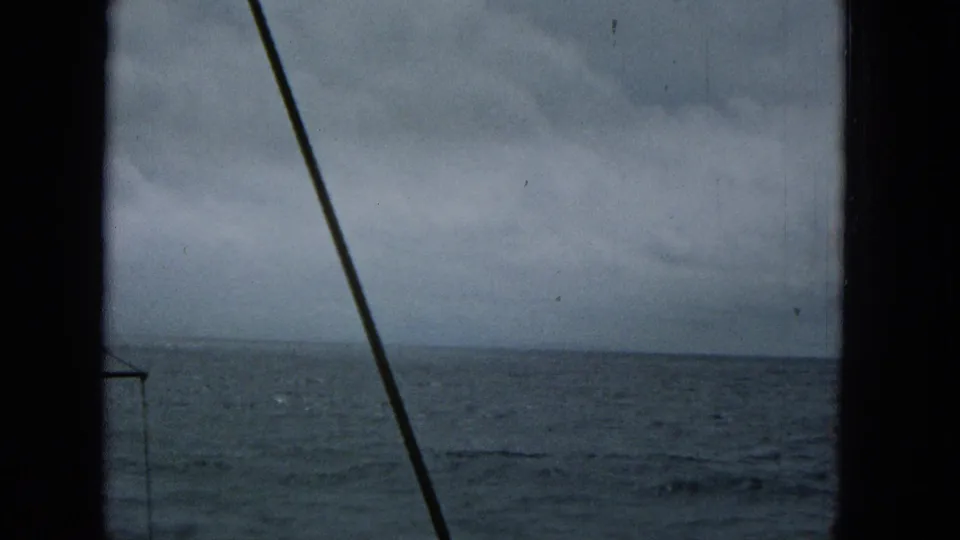 Open ocean stretches under a gray clouded sky as dark boat rigging and edge framing cut across the scanned 8mm home-movie view.
