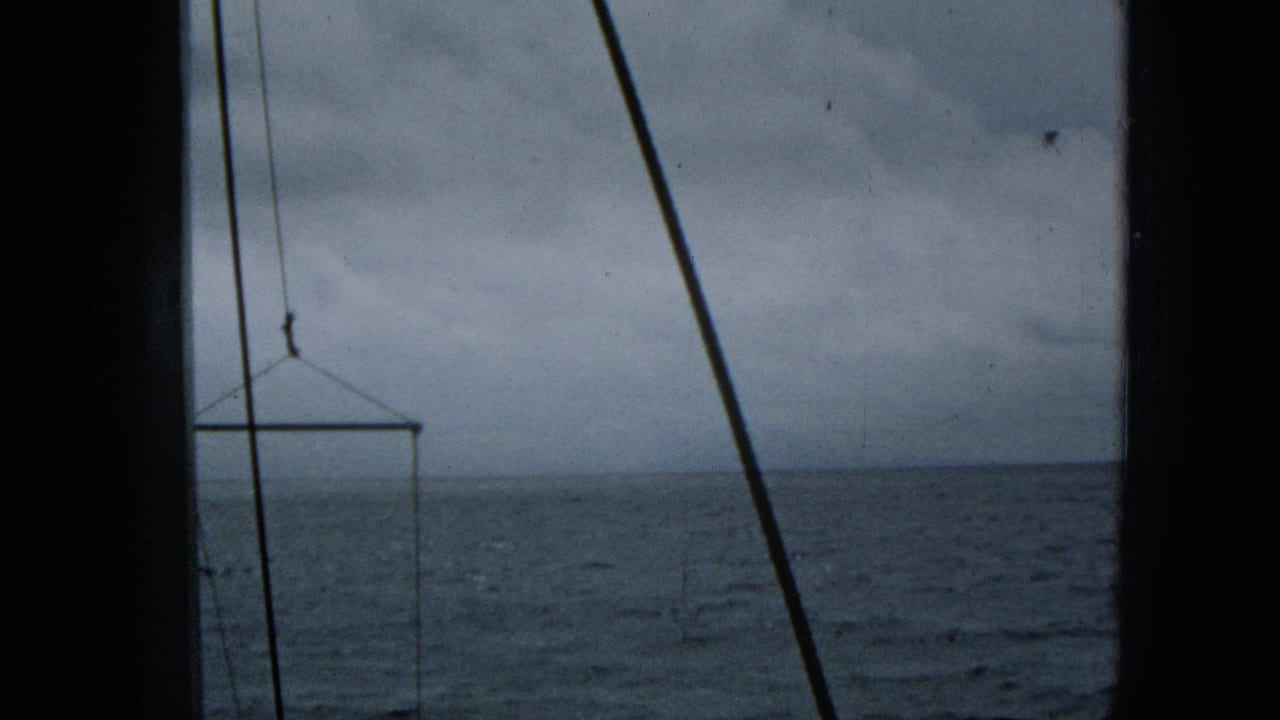 Super 8 frame from the 1965 ocean approach shows blue-gray water, cloudy sky, diagonal rigging, and a suspended rectangular frame near the horizon.