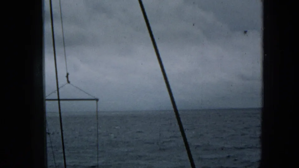 Super 8 frame from the 1965 ocean approach shows blue-gray water, cloudy sky, diagonal rigging, and a suspended rectangular frame near the horizon.