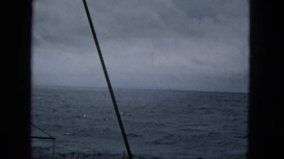 1965 home-movie view from a boat shows rough blue-gray water, a cloudy horizon, dark edge framing, and a diagonal line crossing the scene.