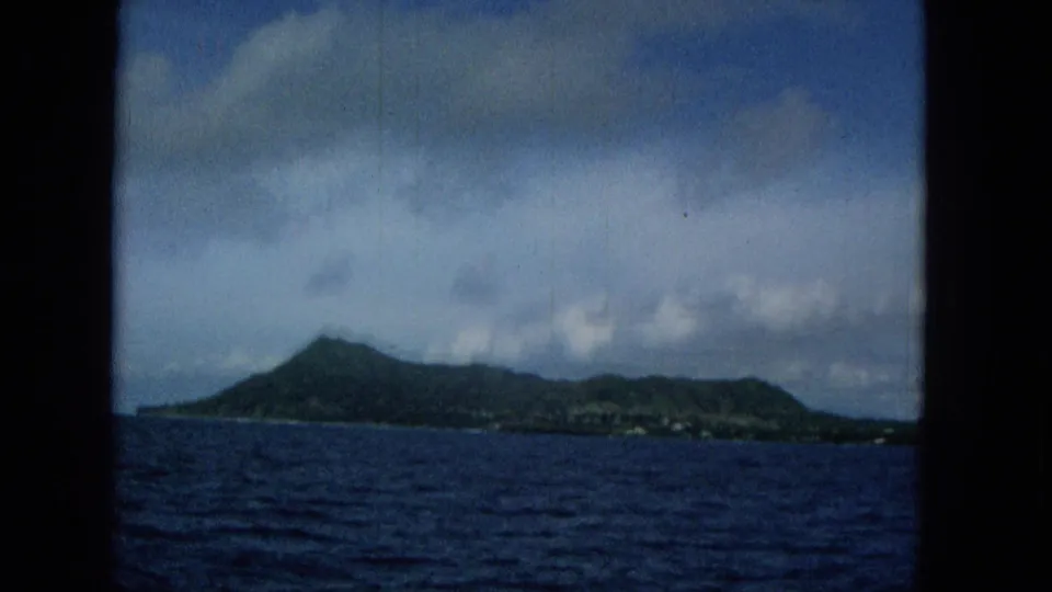 Metadata-supported 1965 view toward Molokai shows a green volcanic island profile across dark blue water, framed by boat structure and clouds.