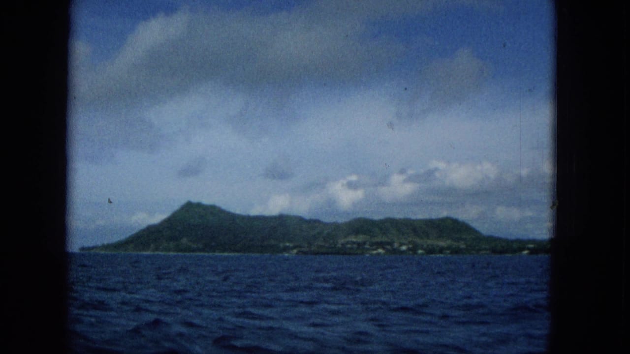 1965 Super 8 boat view of Molokai with a low green ridge, deep blue ocean, partly cloudy sky, and a dark frame edge suggesting a window or porthole.
