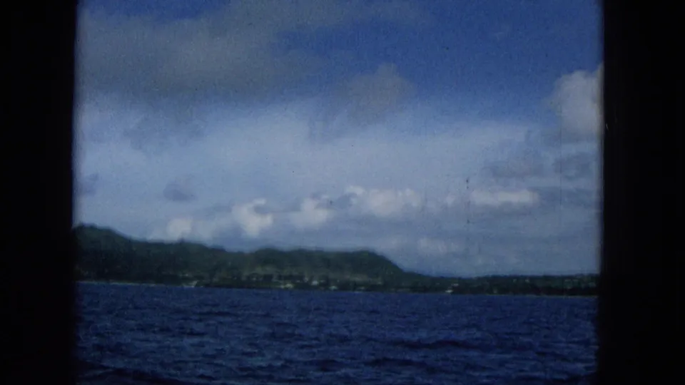 1965 Super 8 frame from a moving boat showing Molokai's low coastline near the left edge, blue water below, and a broad partly cloudy sky overhead.