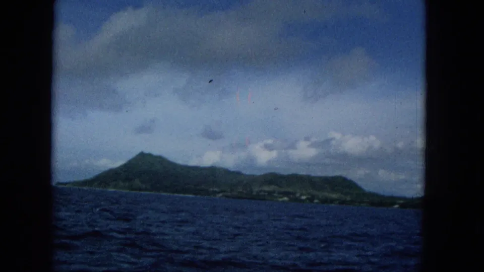 1965 Super 8 view of Molokai from the water, with green mountain slopes, dark blue ocean texture, partly cloudy sky, and visible home-movie frame edges.