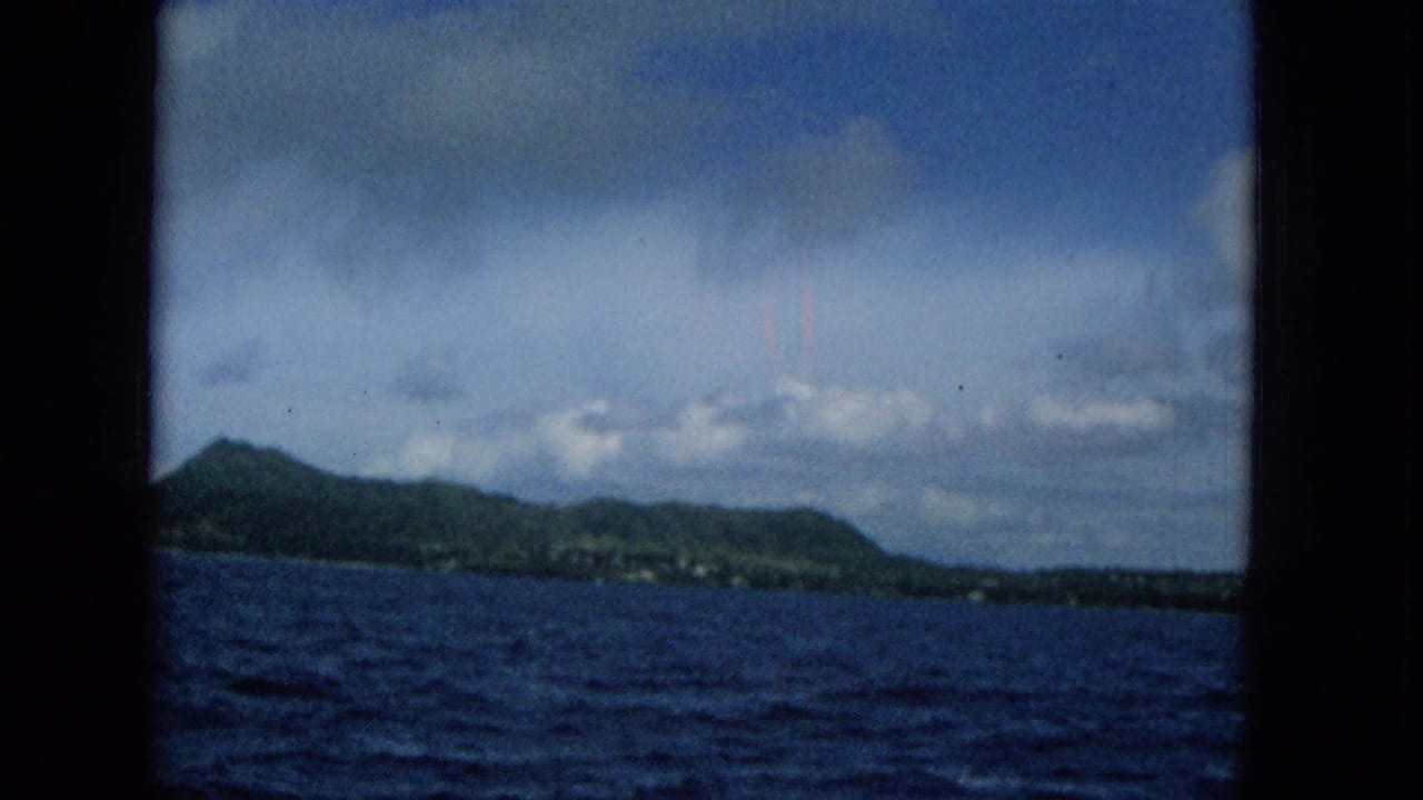 1965 8mm home-movie frame showing a distant Hawaiian island shoreline across deep blue water, with clouds overhead and dark frame edges around the view.