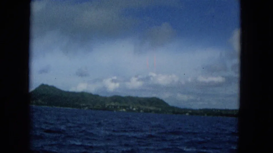 1965 8mm home-movie frame showing a distant Hawaiian island shoreline across deep blue water, with clouds overhead and dark frame edges around the view.