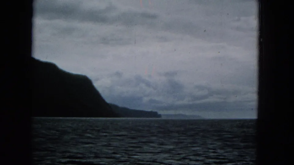 A steep Hawaiian coastline appears as a dark silhouette beyond rippled ocean water, with low clouds and the boat or film frame cropping the scene.