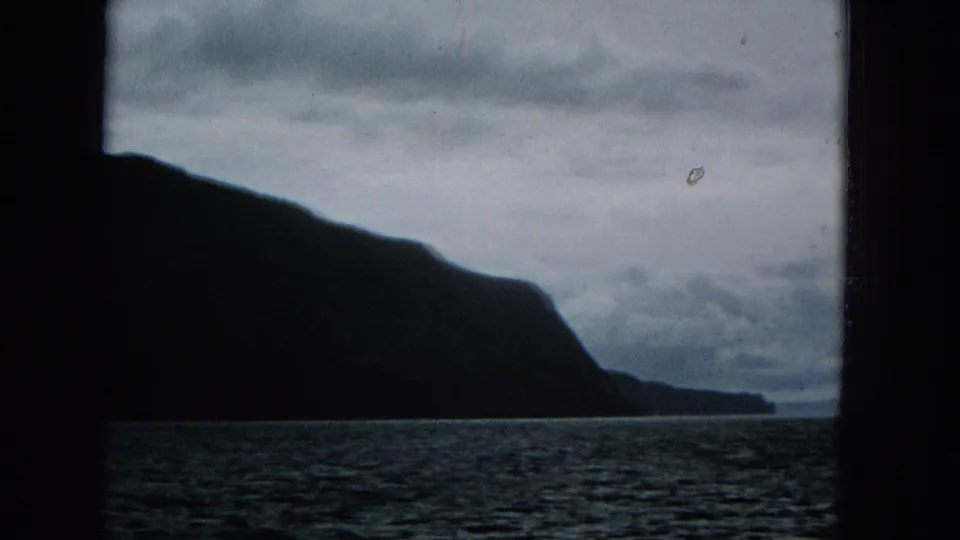 The frame shows a shadowed coastal slope and distant headland across the ocean, with grey clouds and dark structural edges emphasizing the passenger viewpoint.
