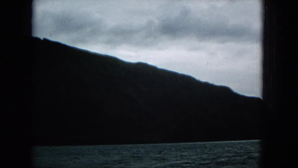 A close, dark silhouette of the island slope fills much of the frame above a thin band of water, with an overcast sky and 8mm color softness.