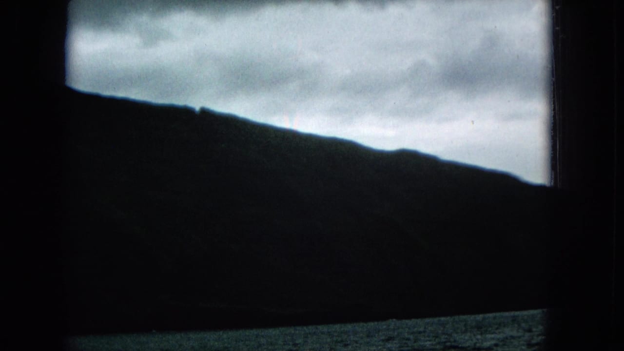 1965 home-movie frame of a silhouetted Molokai mountain ridge and overcast sky seen through a dark vessel window frame.