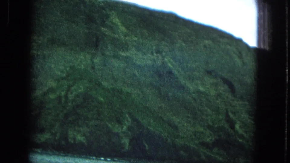 A clearer 1965 boat-view frame of Molokai's green coastal mountain rising above calm water beneath a bright overcast sky.
