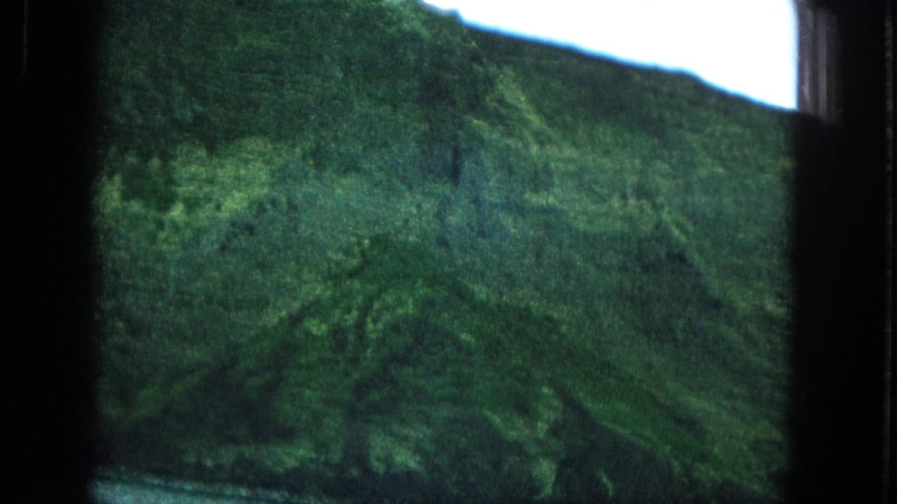 1965 Super 8 frame of a lush green Molokai mountainside viewed through a dark window frame with pale sky above.
