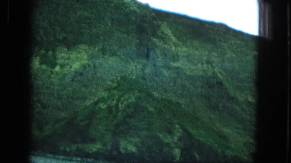 1965 Super 8 frame of a lush green Molokai mountainside viewed through a dark window frame with pale sky above.