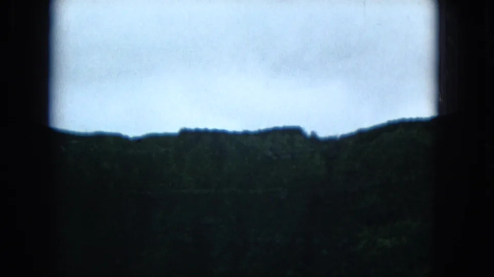 1965 Super 8 frame of a dark Molokai ridge line beneath a pale overcast sky, seen through the right side of a window frame.