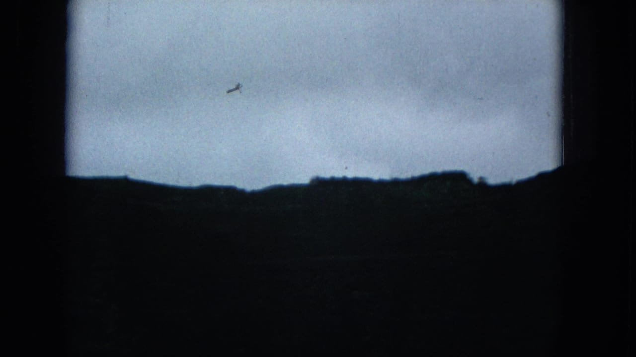 1965 8mm home-movie frame showing a dark, distant hill silhouette under a pale overcast sky, with visible grain and black frame edges.