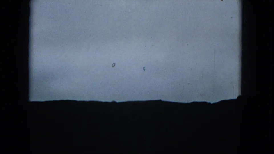 A low, dark ridgeline sits beneath a washed grey sky in this 1965 8mm frame, with strong edge shadow and visible film texture.
