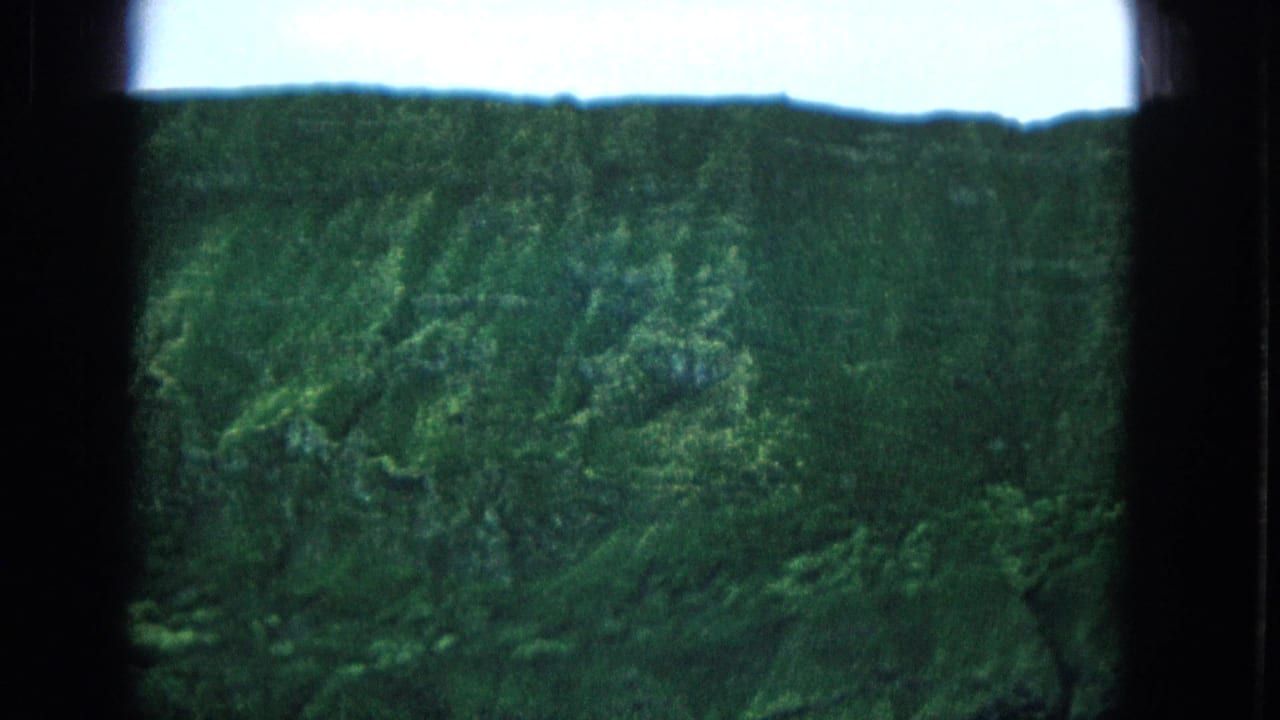 1965 Super 8 frame from Molokai, Hawaii, showing distant green cliff slopes under a pale sky with dark film gate edges visible.
