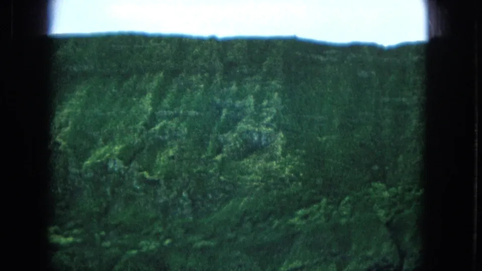 1965 Super 8 frame from Molokai, Hawaii, showing distant green cliff slopes under a pale sky with dark film gate edges visible.
