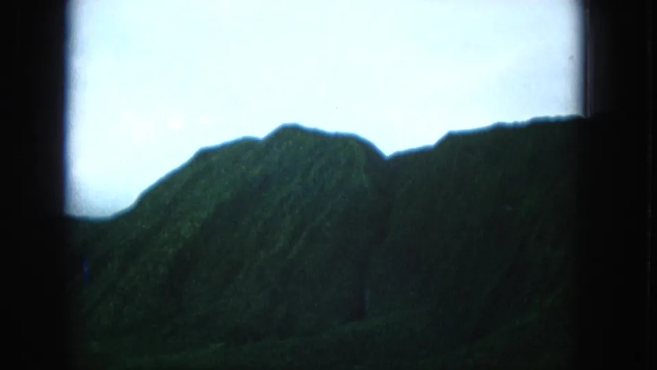 1965 Super 8 frame from Molokai, Hawaii, showing a dark green ridgeline against a bright overcast sky within the film frame.
