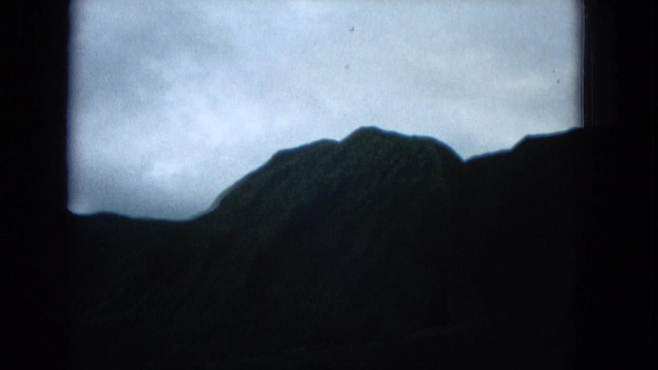 1965 home-movie frame of a dark Molokai mountain ridge silhouetted against a cloudy blue-gray sky.