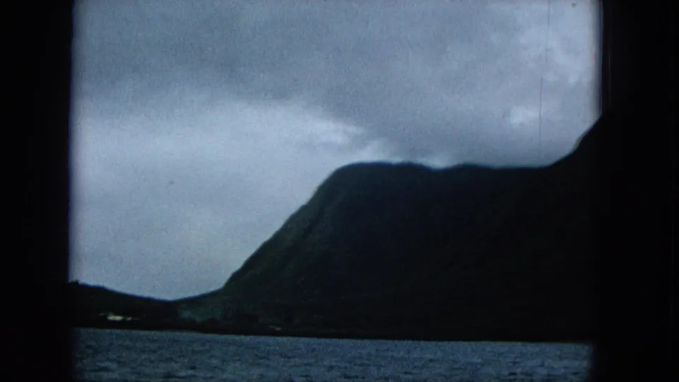 1965 home-movie view of a shadowed Molokai mountain rising beyond ocean water and a low shoreline.