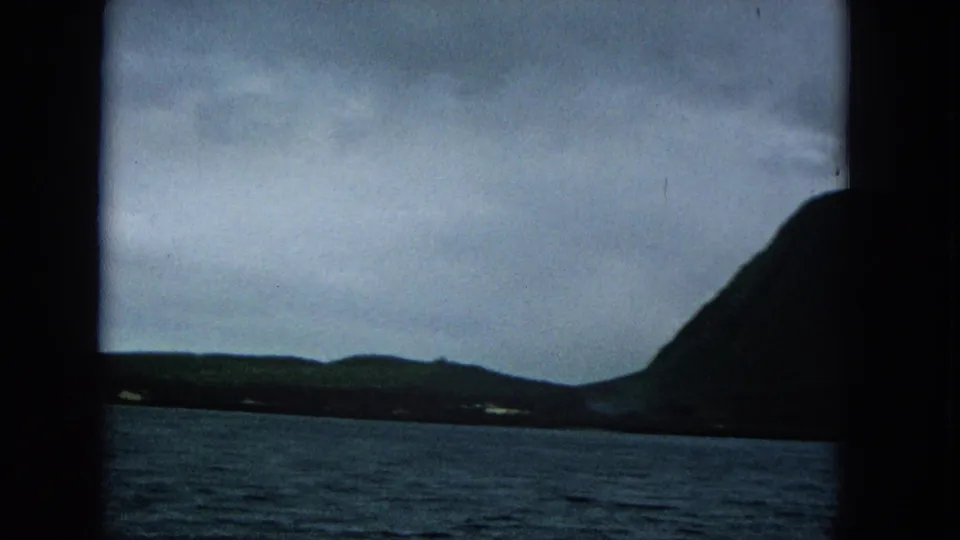 1965 Molokai coastline frame showing open water, distant low land, and a dark mountain slope at the right edge.