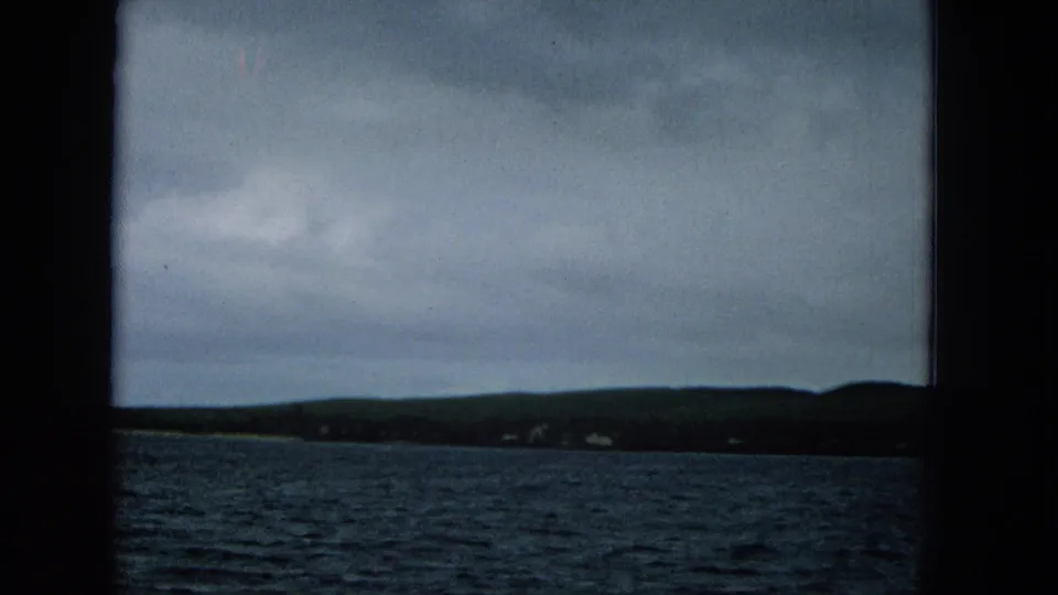 Boat-view Super 8 frame of a low Molokai shoreline, overcast sky, and dark blue water framed by the film gate.