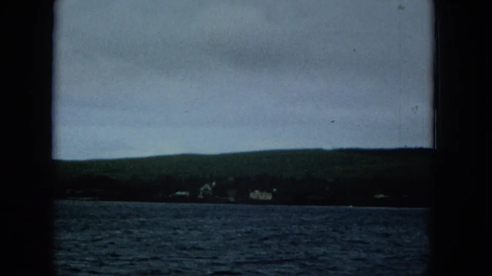 1965 Molokai home-movie view of small shoreline structures below green hills, with rippled water in the foreground.